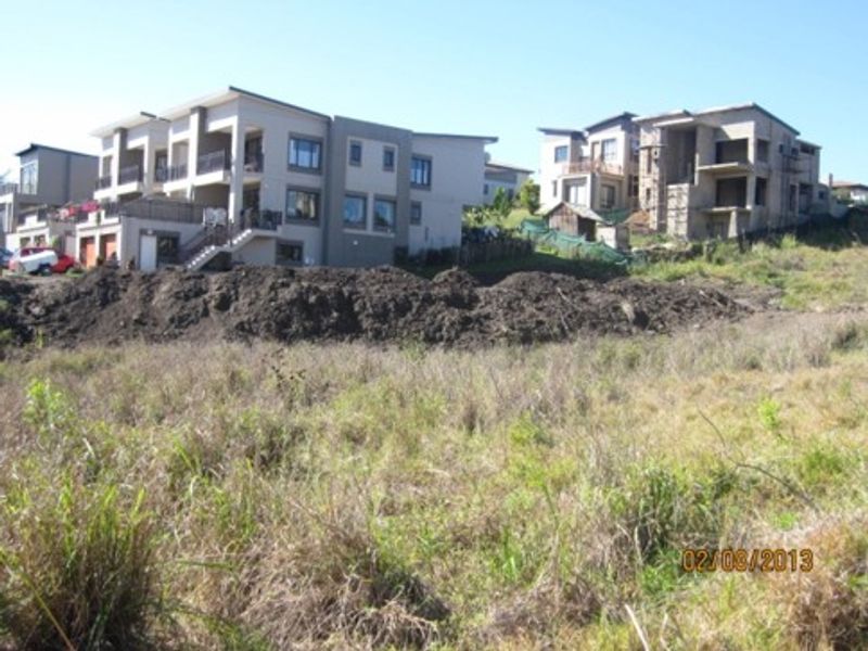 A view of a vacant residential plot with sloped terrain, situated in a developed neighborhood with surrounding multi-story residential properties.