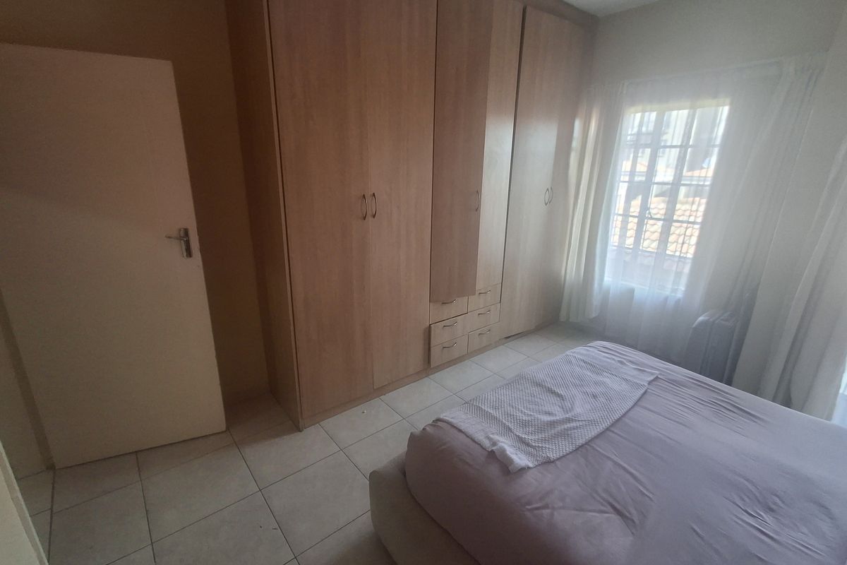 A bedroom featuring built-in wooden cupboards, tiled flooring, and a window with security bars, offering a functional and neutral living space.