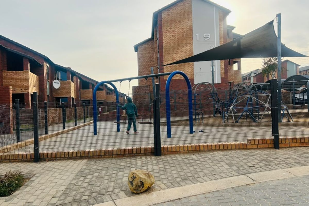 Communal outdoor area featuring a playground with shade sail, enclosed by perimeter fencing, situated between residential apartment blocks.