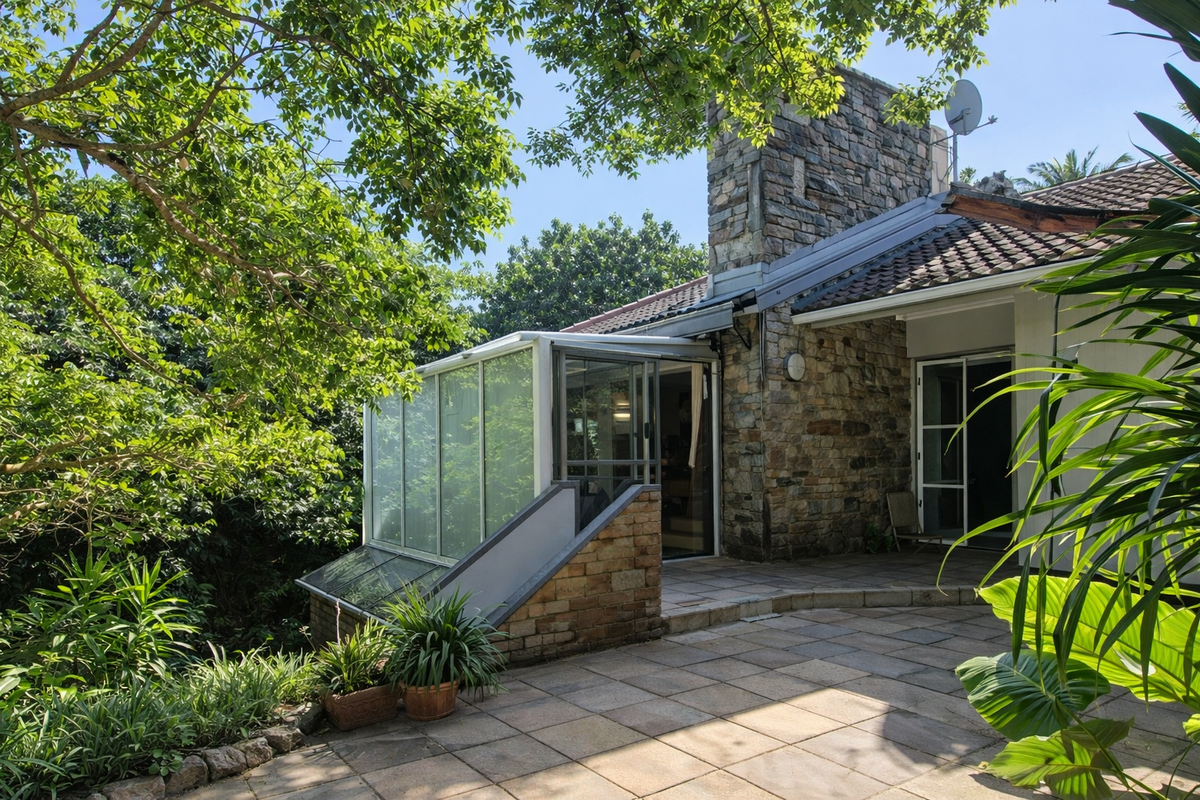 Rear exterior view featuring a stone-clad chimney, a paved patio area, and a glass-enclosed conservatory extension surrounded by mature garden foliage.