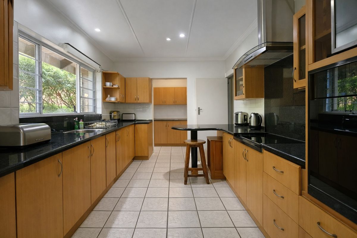 A functional kitchen featuring wooden cabinetry, granite countertops, built-in oven, stainless steel extractor fan, and tiled flooring with an adjacent scullery area.