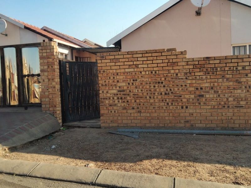 Front exterior view of a cluster property featuring a face brick boundary wall, metal security gate, and paved driveway area.