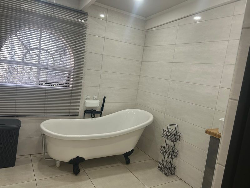 A clean bathroom featuring a classic freestanding bathtub with claw feet, neutral wall tiling, and a window with venetian blinds.