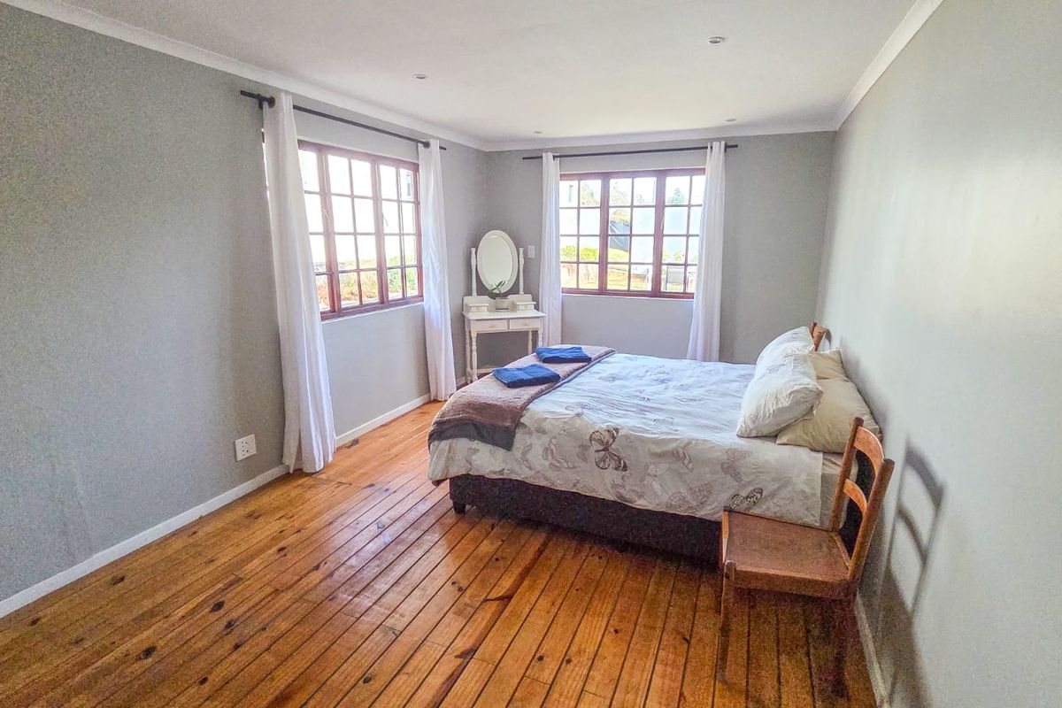 A bright and spacious bedroom featuring warm wooden flooring, neutral wall tones, and large timber-framed windows that provide ample natural light.