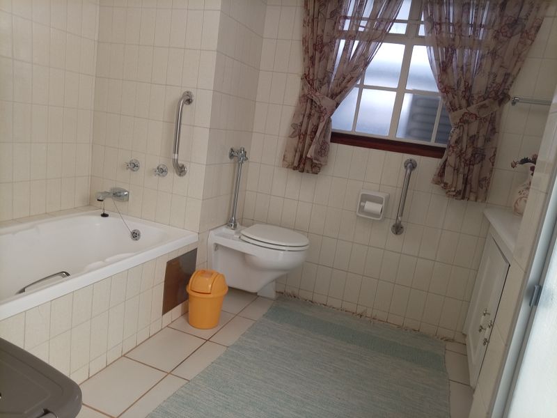 A functional bathroom featuring a built-in bathtub, wall-mounted toilet, vanity cabinet, and safety grab rails, finished with neutral wall and floor tiling.