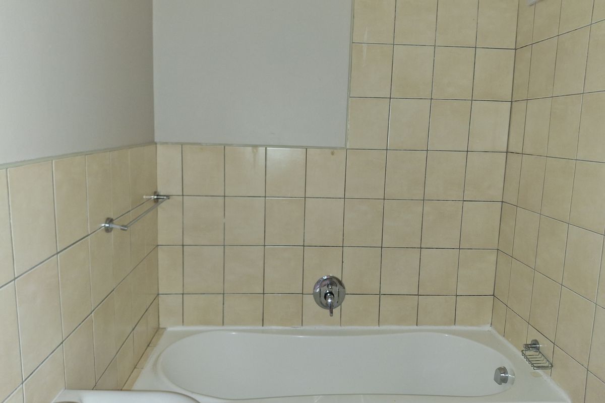 A dated bathroom featuring a built-in bathtub with wall-mounted shower mixer and beige ceramic wall tiling.