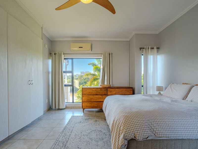 A bright bedroom featuring tiled flooring, built-in cupboards, a ceiling fan, and a wall-mounted air conditioning unit with views of the surrounding greenery.
