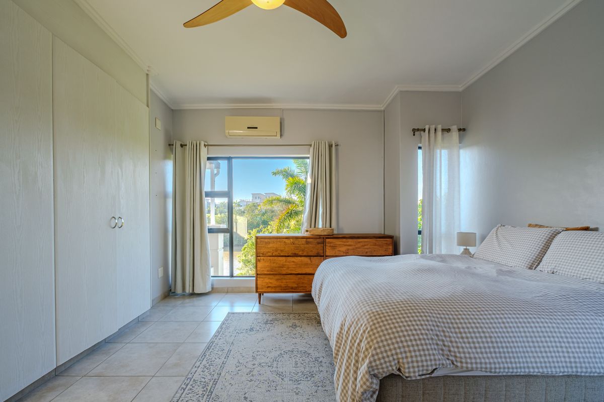 A bright bedroom featuring tiled flooring, built-in cupboards, a ceiling fan, and a wall-mounted air conditioning unit with views of the surrounding greenery.