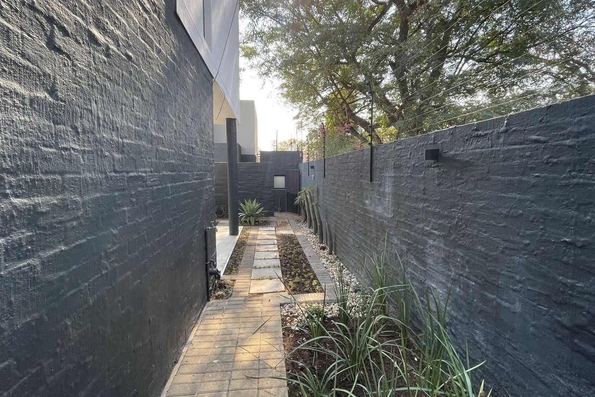 A side walkway featuring a paved path, landscaped garden beds, and a high painted brick boundary wall with integrated electric fencing.