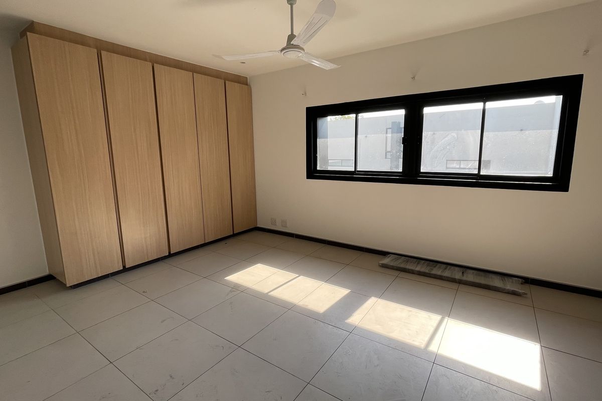 A bright, empty bedroom featuring neutral tiled flooring, ample built-in wooden cupboards, a ceiling fan, and a large sliding window.