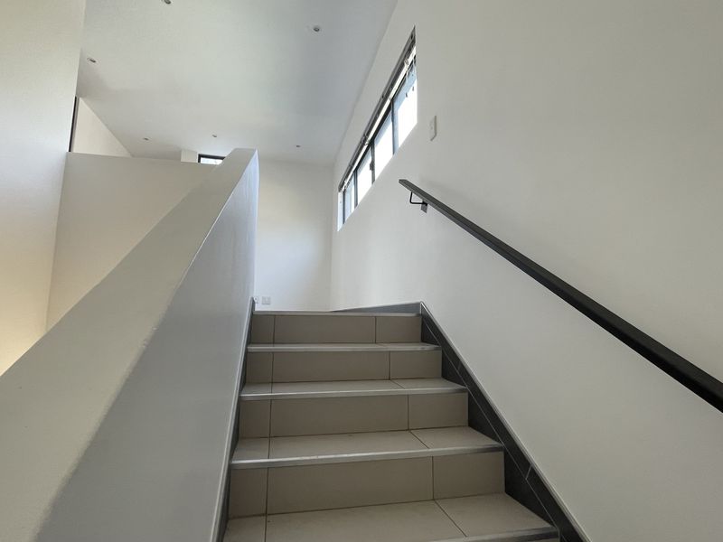 A bright, modern staircase featuring tiled steps, a sleek metal handrail, and recessed ceiling lighting, leading to an upper level.