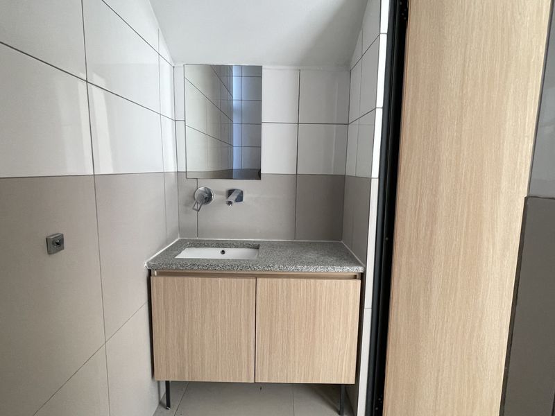 A modern, compact bathroom featuring a vanity cabinet with a stone countertop, undermount sink, wall-mounted faucet, and neutral ceramic wall tiling.