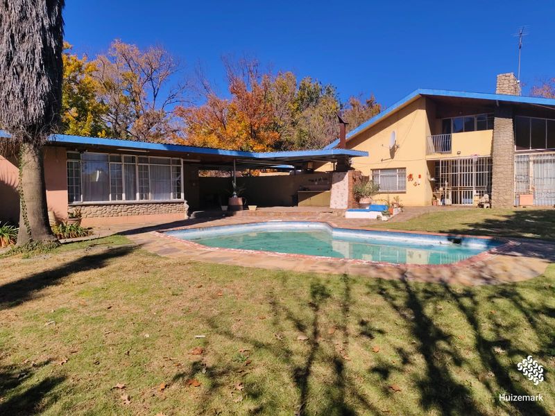 Rear exterior view featuring a swimming pool, covered patio area, stone chimney, and established garden setting.