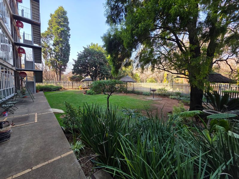 A spacious rear garden featuring a concrete patio, well-maintained lawn, mature trees, and thatched gazebos, providing a peaceful outdoor area for the flat.
