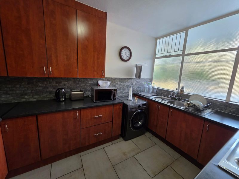 A functional kitchen featuring wood-finish cabinetry, dark countertops, a double sink, and tiled flooring with ample natural light from a large window.