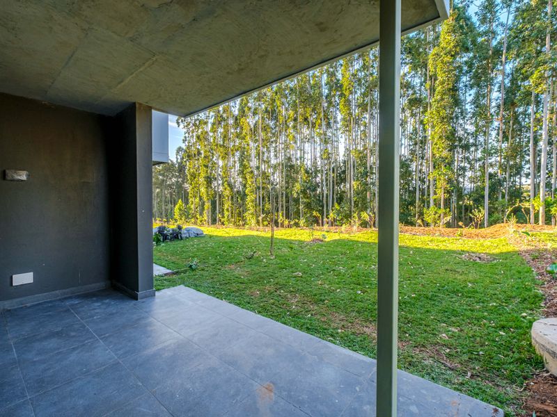 A covered patio area featuring tiled flooring and a concrete ceiling, overlooking a private garden with a scenic treed backdrop.