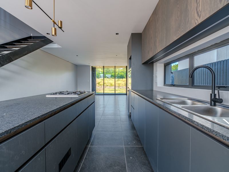 Modern kitchen featuring dark cabinetry, stone countertops, integrated gas hob, and sleek finishes, leading to an open-plan living area with staircase access.