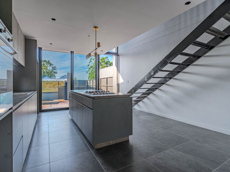 Modern open-plan kitchen featuring a central island with gas cooktop, built-in oven, sleek cabinetry, and a striking metal staircase, all bathed in natural light.