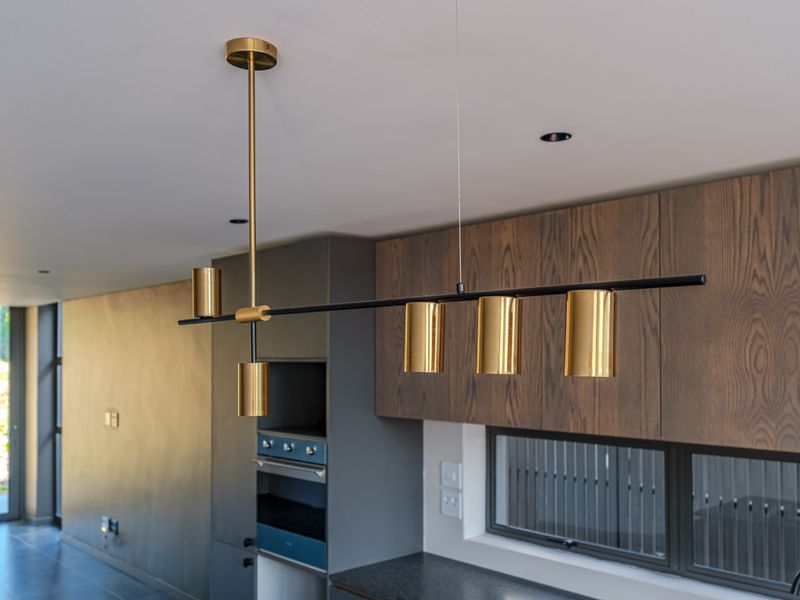 Modern kitchen featuring dark wood cabinetry, built-in oven, stone countertops, and contemporary gold pendant lighting in an open-plan layout.