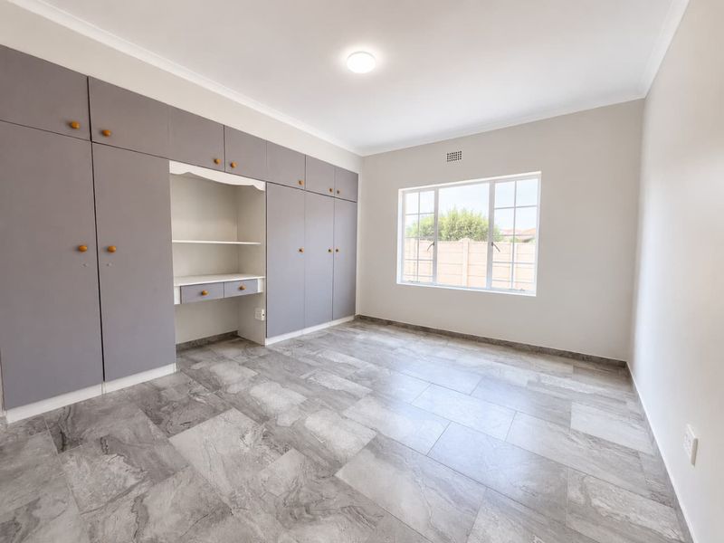 A bright, empty bedroom featuring modern grey tiled flooring, extensive built-in cupboards with an integrated desk area, and a large window.