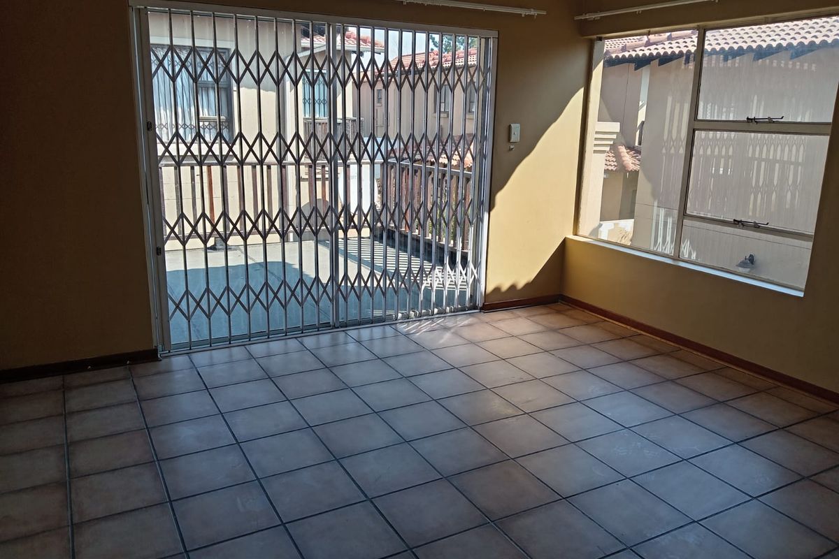 An empty living room featuring tiled flooring, large windows, and a sliding glass door with a security gate leading to an outdoor area.