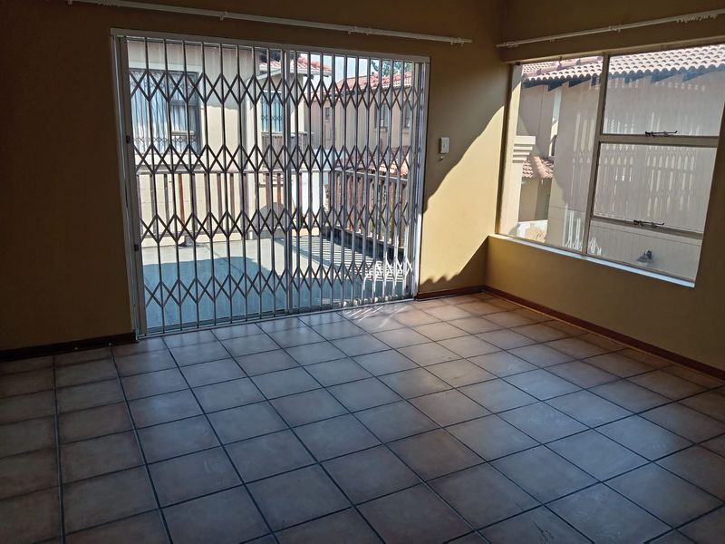 An empty living room featuring tiled flooring, large windows, and a sliding glass door with a security gate leading to an outdoor area.