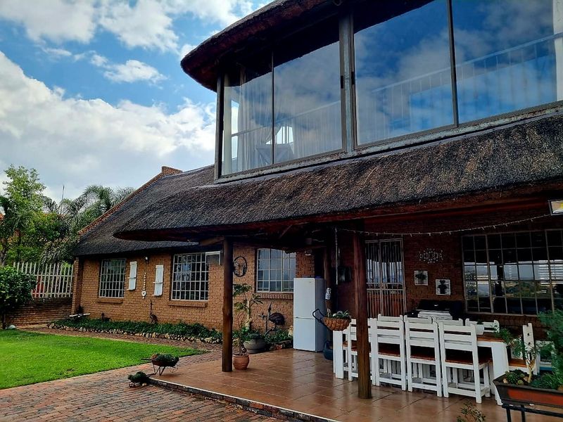Rear view of a double-story house featuring a traditional thatched roof, brick exterior, and a spacious tiled patio area with large upper-level glass windows.