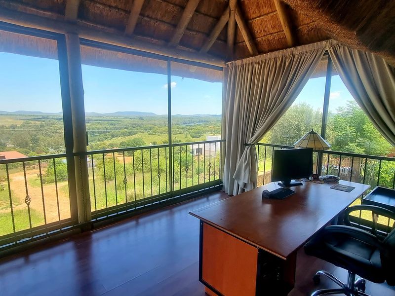 A bright home office featuring a traditional thatch roof, polished timber flooring, and expansive glass doors offering panoramic views of the surrounding landscape.