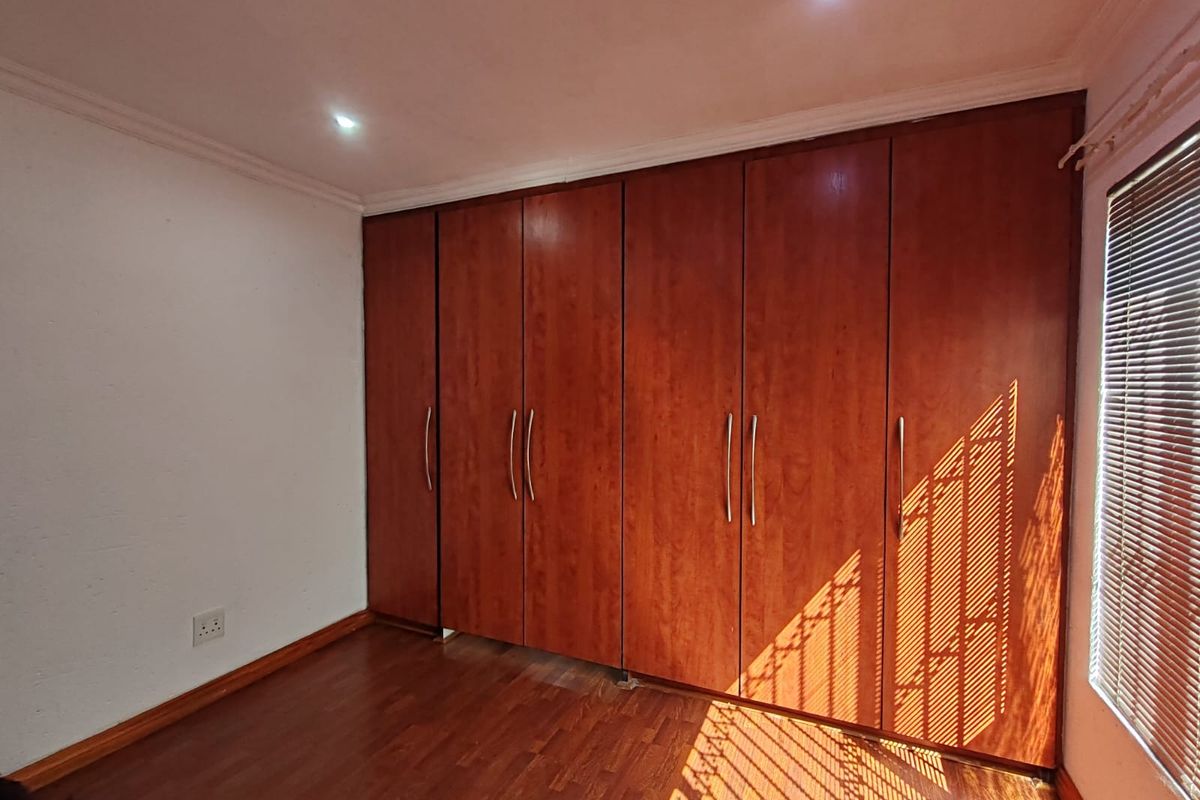 A clean bedroom featuring extensive floor-to-ceiling built-in wooden cupboards, warm laminate flooring, and venetian blinds allowing natural light.