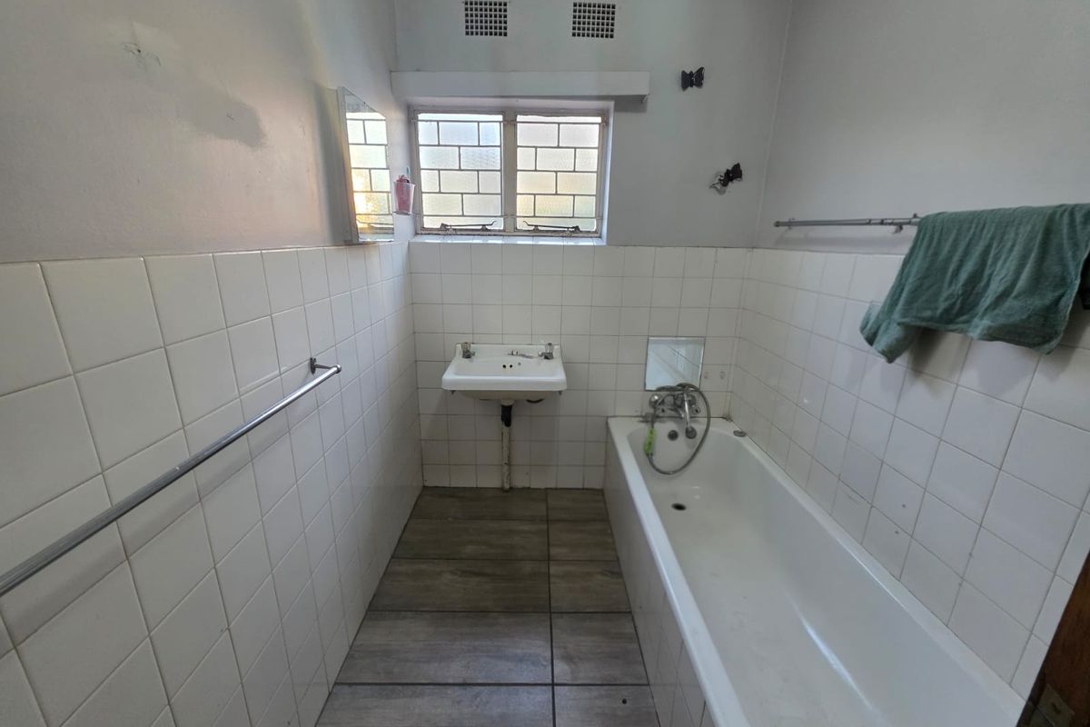 A dated bathroom featuring a built-in bathtub, wall-mounted basin, and tiled finishes, requiring cosmetic updates and repairs to wall fixtures.