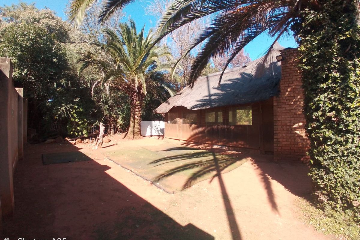 Rear garden area featuring a thatched roof structure with a brick chimney and artificial lawn, surrounded by mature trees and boundary walls.