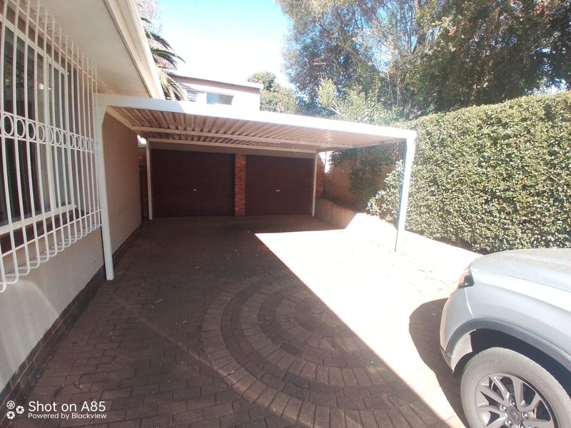 A double garage with roller shutter doors and an attached carport, featuring a paved driveway and security bars on the adjacent building windows.