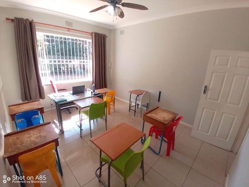 A functional office space featuring tiled flooring, a ceiling fan, and large windows with security bars, currently set up as a study or classroom area.