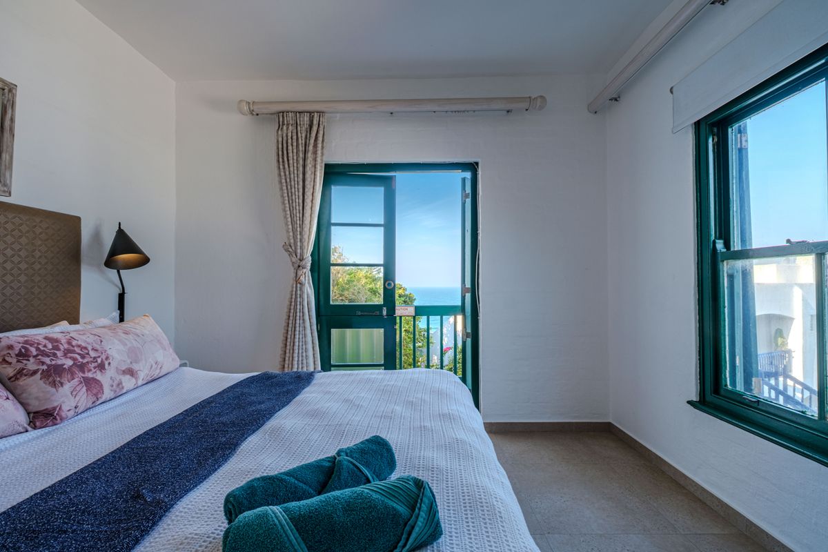 A bright bedroom featuring tiled floors, a wall-mounted reading light, and french doors opening to a balcony with scenic ocean views.