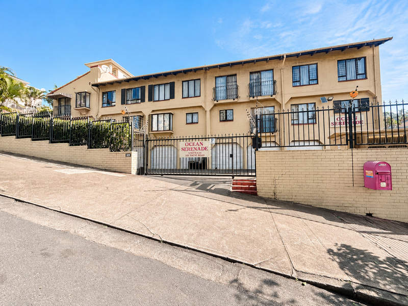 Front elevation of Ocean Serenade apartment complex featuring a secure gated entrance, concrete driveway, and multi-level residential units with balconies.