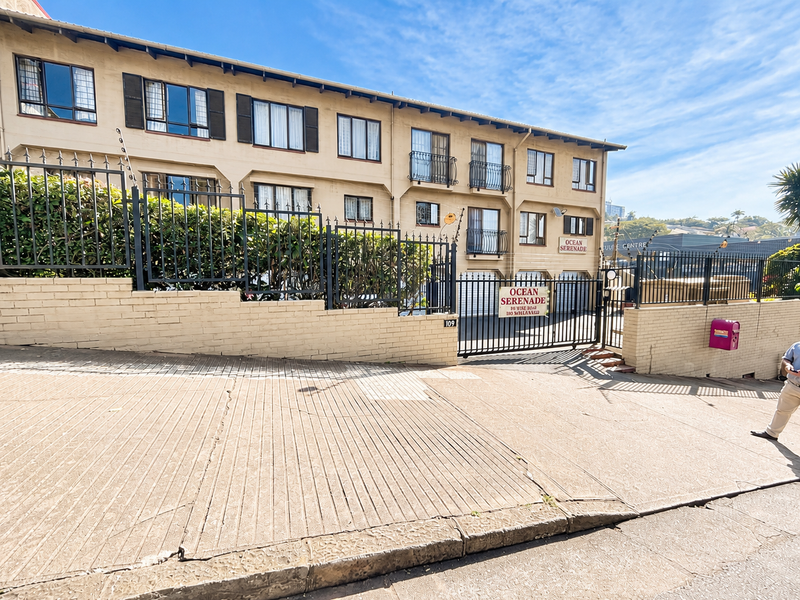 Front exterior view of Ocean Serenade apartment complex featuring a gated entrance, concrete driveway, and multi-level residential units with balconies.