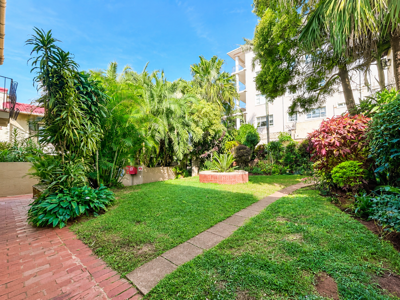 A lush, well-maintained communal garden area featuring mature tropical landscaping, brick pathways, and a central brick planter box within an apartment complex.