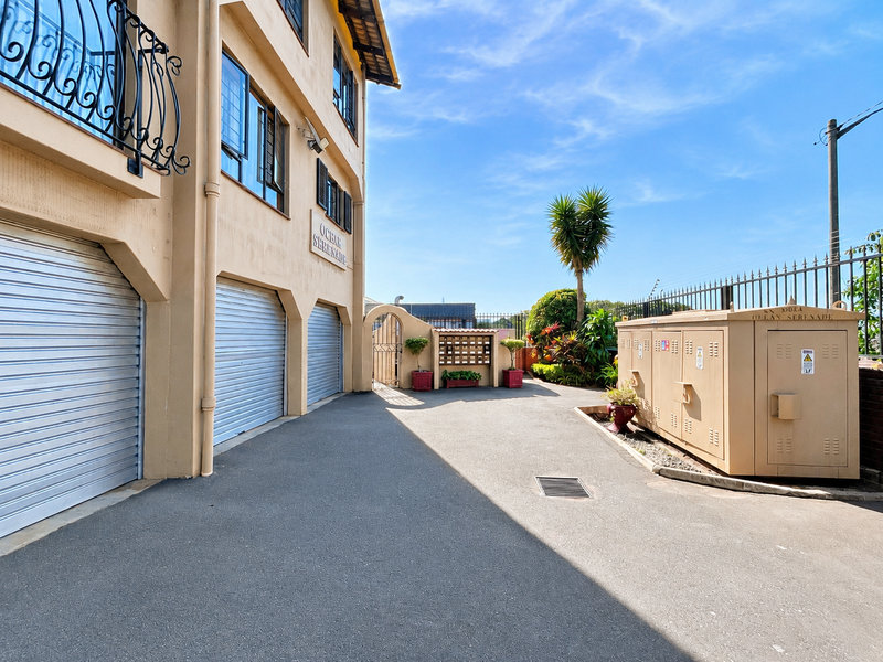 Exterior view of the apartment complex featuring multiple roller shutter garage doors, paved driveway access, and secure perimeter fencing.