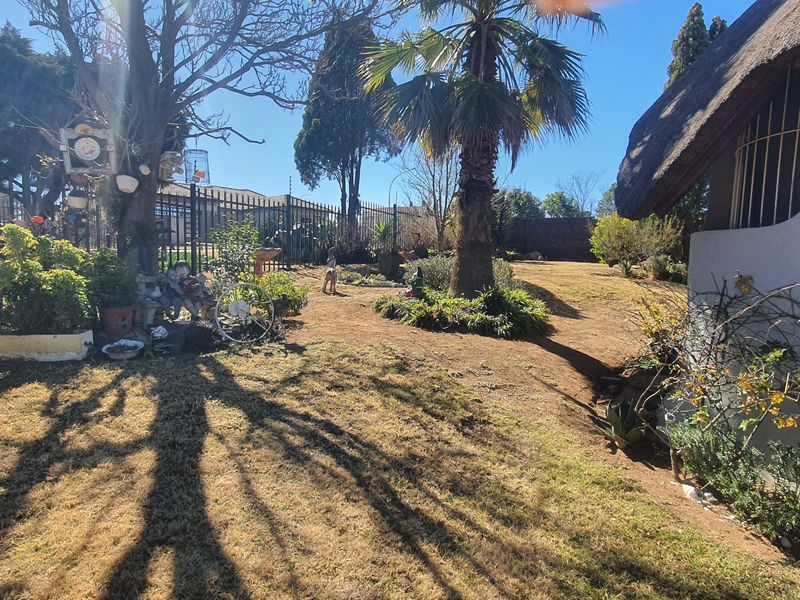 A spacious garden area featuring mature trees, established landscaping, and metal perimeter fencing, adjacent to a property with a traditional thatched roof.