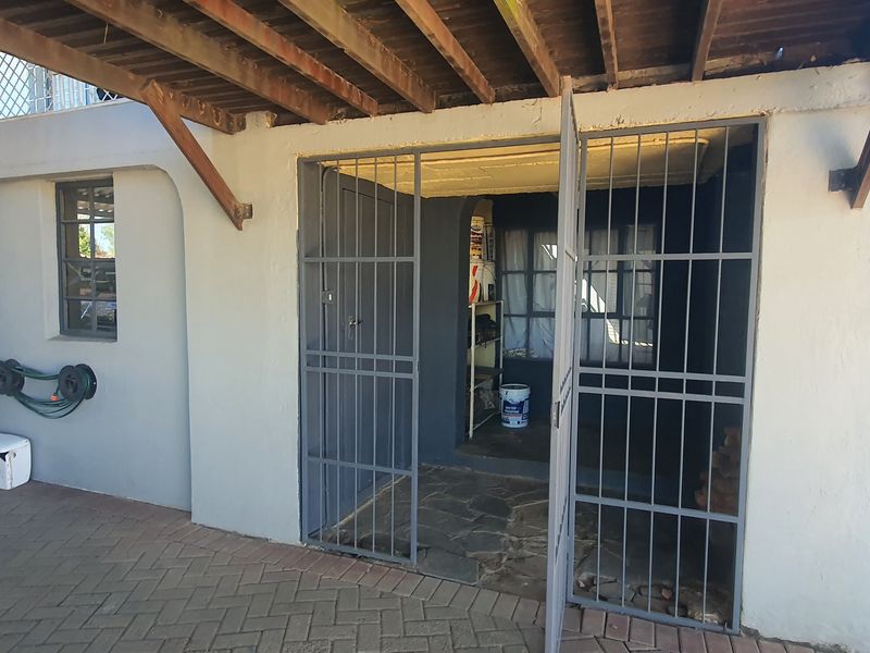 A utility storage area located beneath a timber deck, featuring a secure metal gate and stone flooring, accessible from the paved driveway.