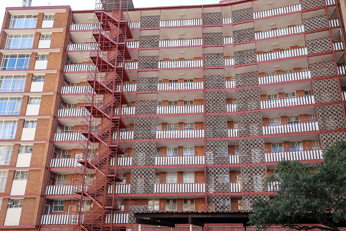 Exterior view of a multi-story apartment building featuring a prominent red metal fire escape, decorative breeze block screens, and multiple private balconies.