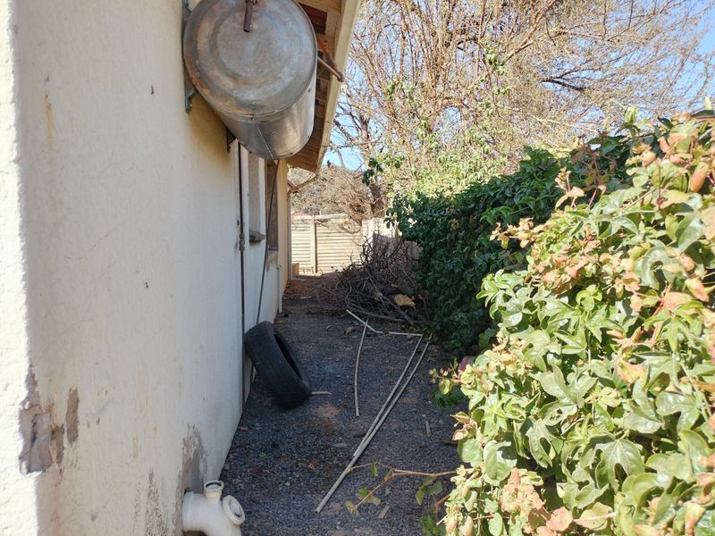 Rear exterior side passage showing an external geyser, exposed plumbing, and unmaintained garden area.