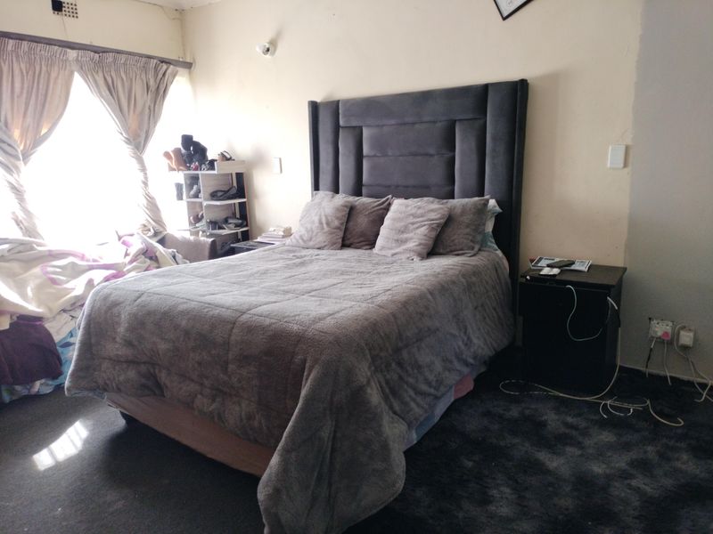 A bedroom featuring a large upholstered headboard, dark carpeting, and natural light from a window with curtains. The walls show signs of wear.