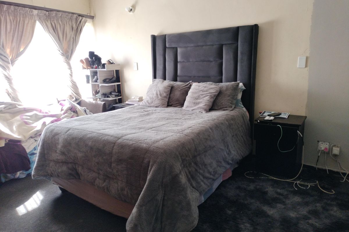 A bedroom featuring a large upholstered headboard, dark carpeting, and natural light from a window with curtains. The walls show signs of wear.