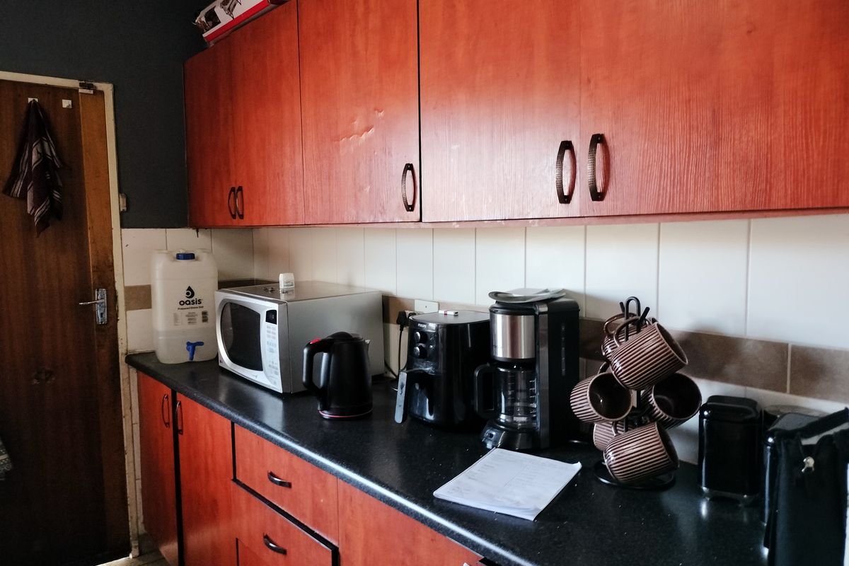 A functional kitchen area featuring wood-look laminate cabinetry, dark countertops, and tiled splashbacks, showing signs of wear and dated finishes.