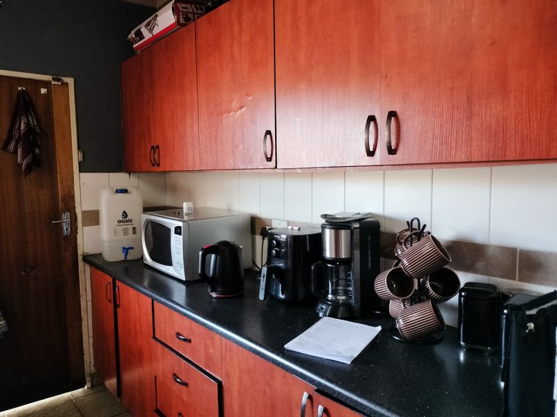 A functional kitchen area featuring wood-look laminate cabinetry, dark countertops, and tiled splashbacks, showing signs of wear and dated finishes.