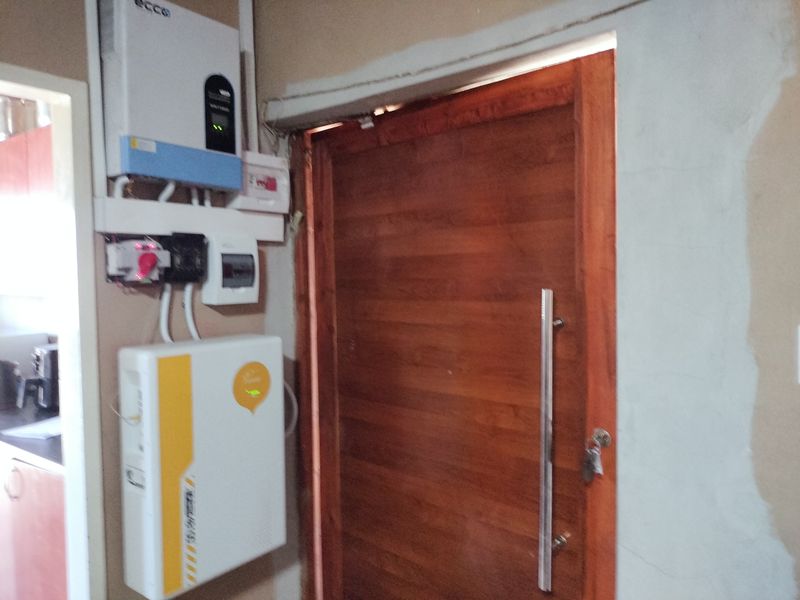 Utility area featuring a solar inverter and battery storage system installed next to a wooden door, with unfinished wall surfaces.