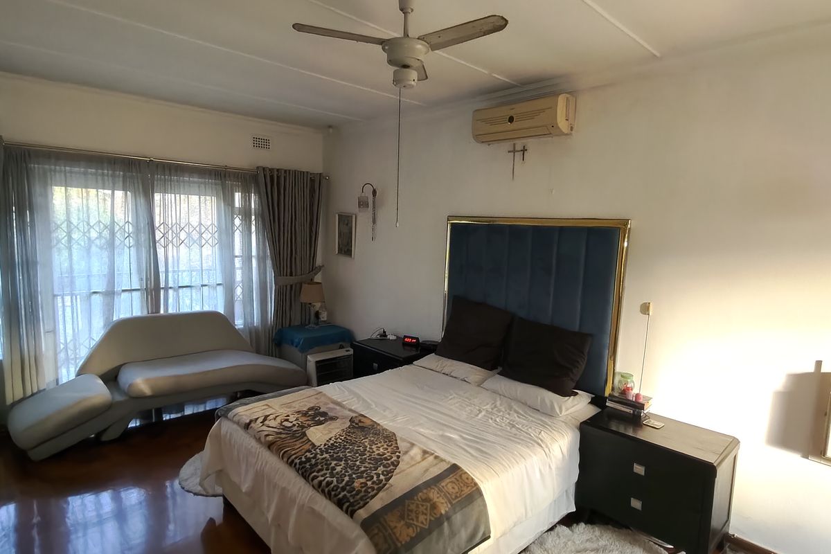 A bedroom featuring hardwood flooring, a ceiling fan, and a wall-mounted air conditioning unit, with large windows secured by metal security gates.