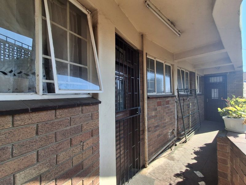 A covered exterior walkway or balcony area featuring brick facade, security gates, and paved flooring, showing signs of age and weathering.