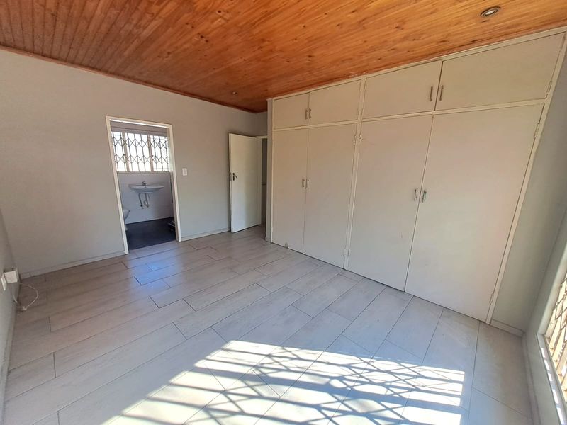 A spacious bedroom featuring built-in cupboards, a warm wooden ceiling, and tiled flooring, with an adjoining en-suite bathroom.